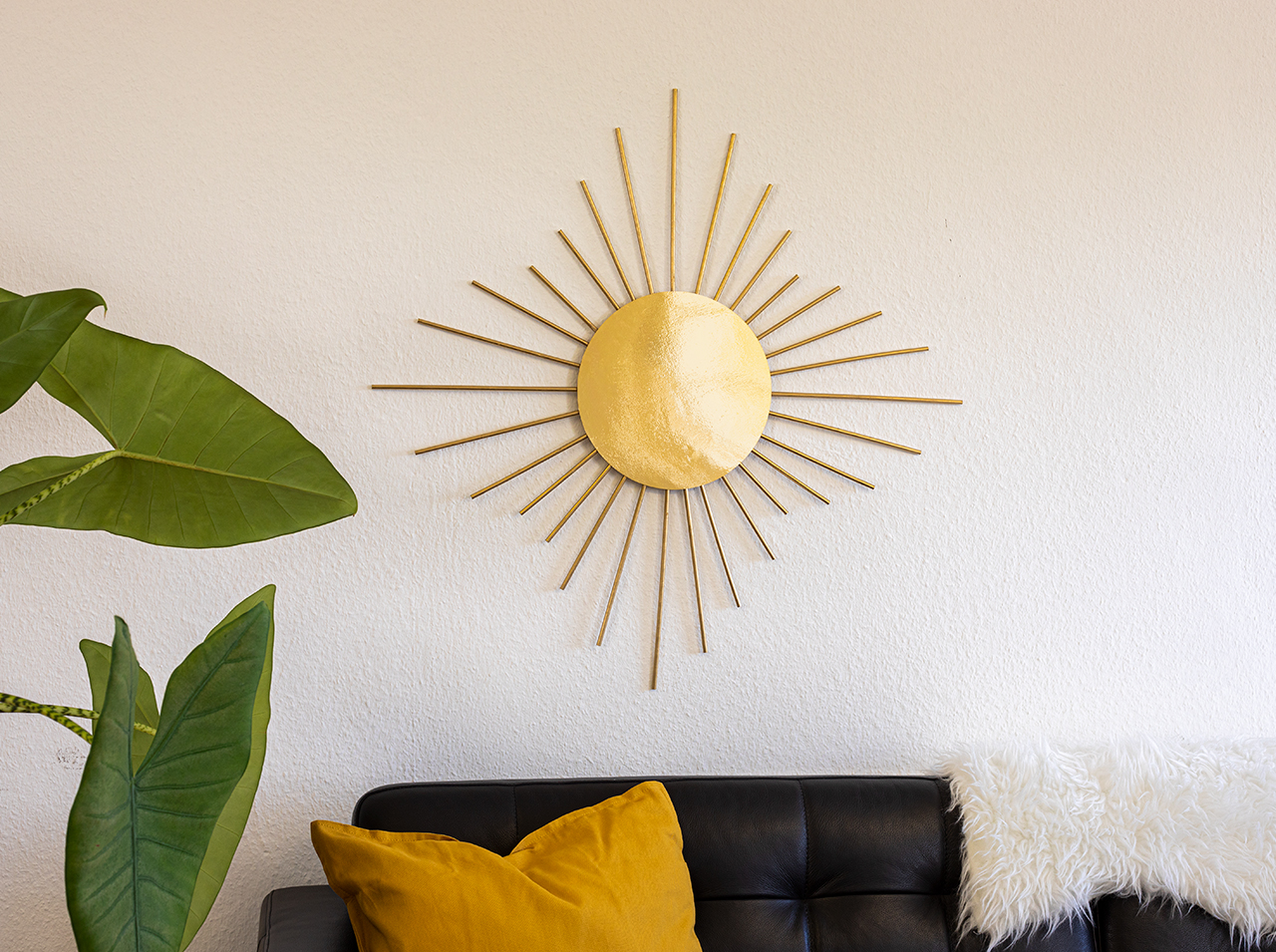 Sur un mur blanc est accroché un miroir en forme de soleil avec un cercle de film doré à effet miroir et des tiges en bois dorées tout autour.